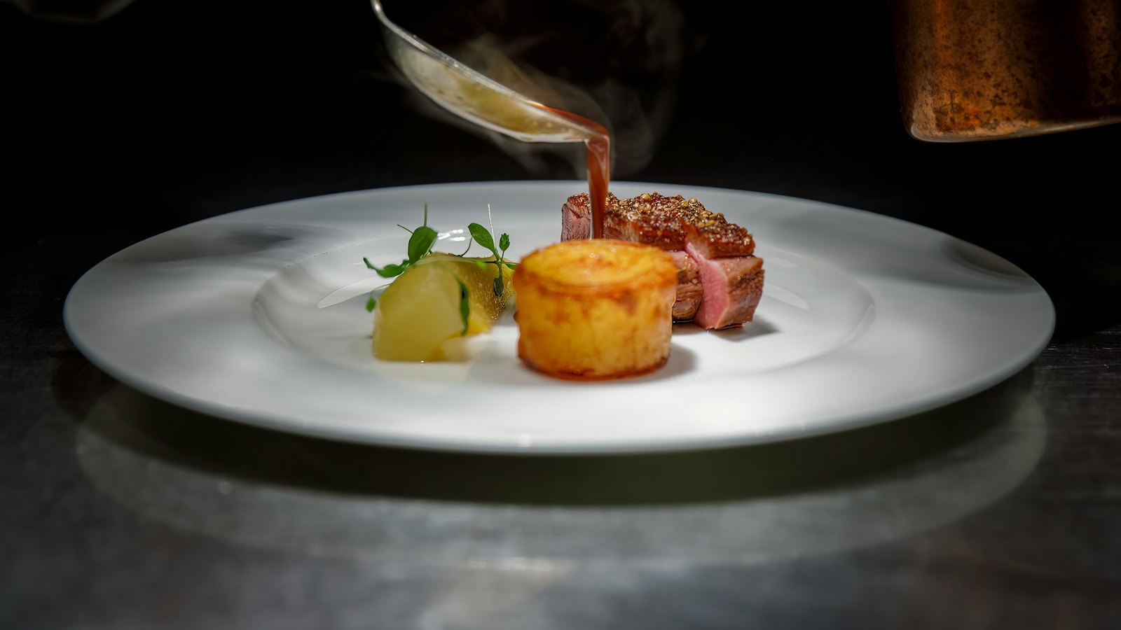 A chef pours sauce over a plated dish of duck and potato on a white plate, lit by a single spotlight against a dark kitchen