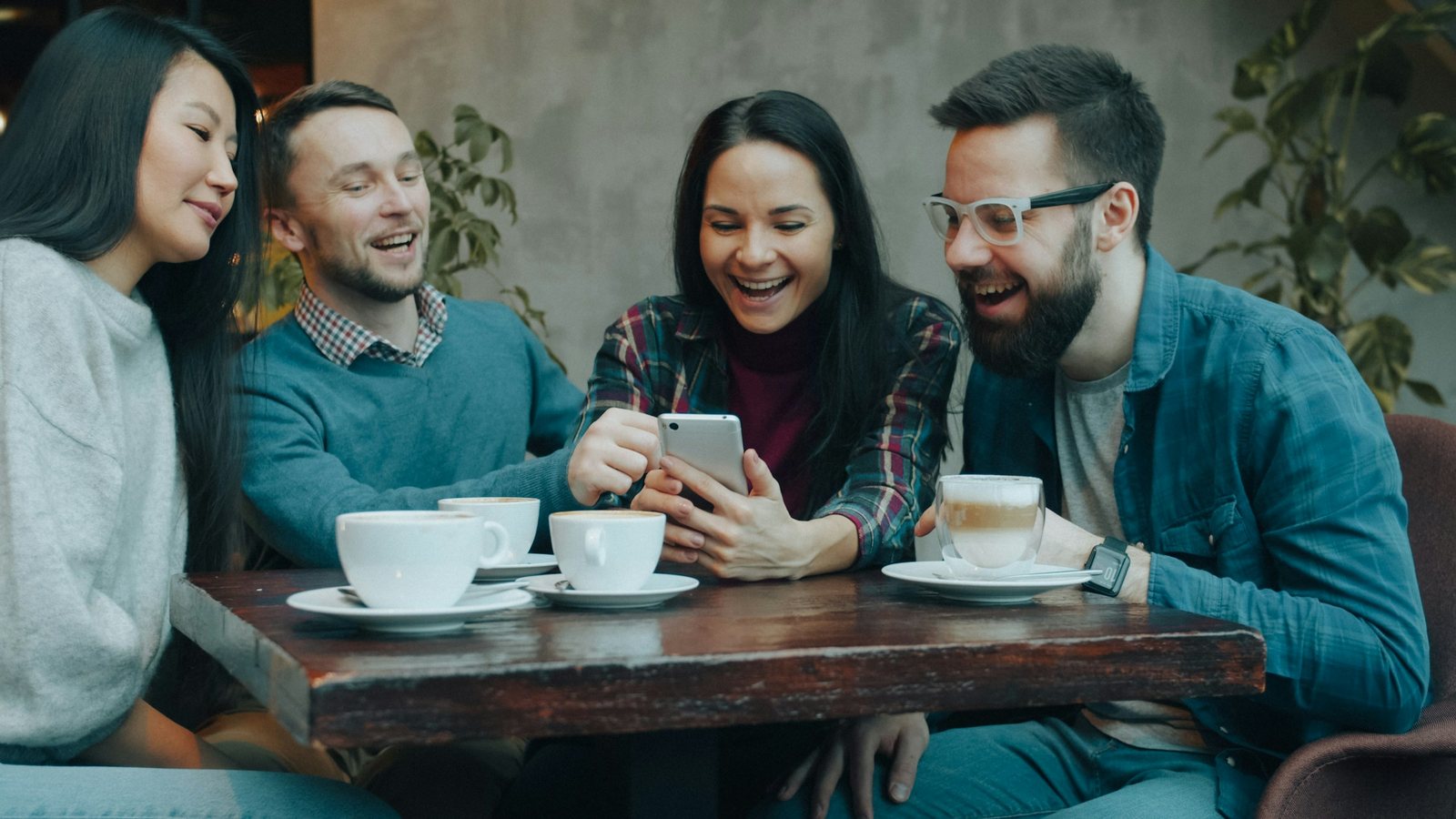 Four friends around a café table with coffee cups, laughing together at something on one of their phones