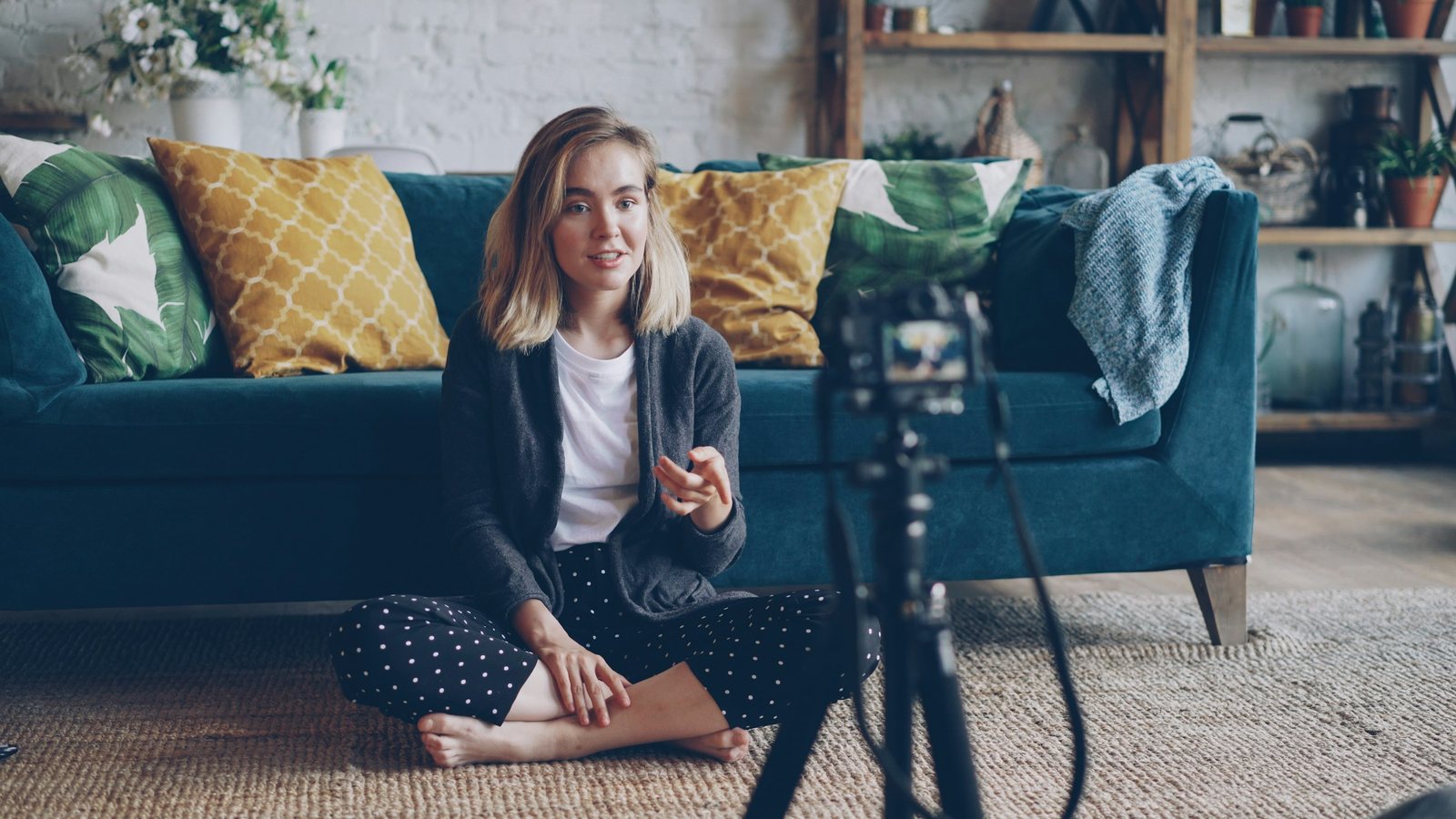 A young woman sits cross-legged on the floor of a bright living room, talking to a camera on a tripod in front of a teal sofa