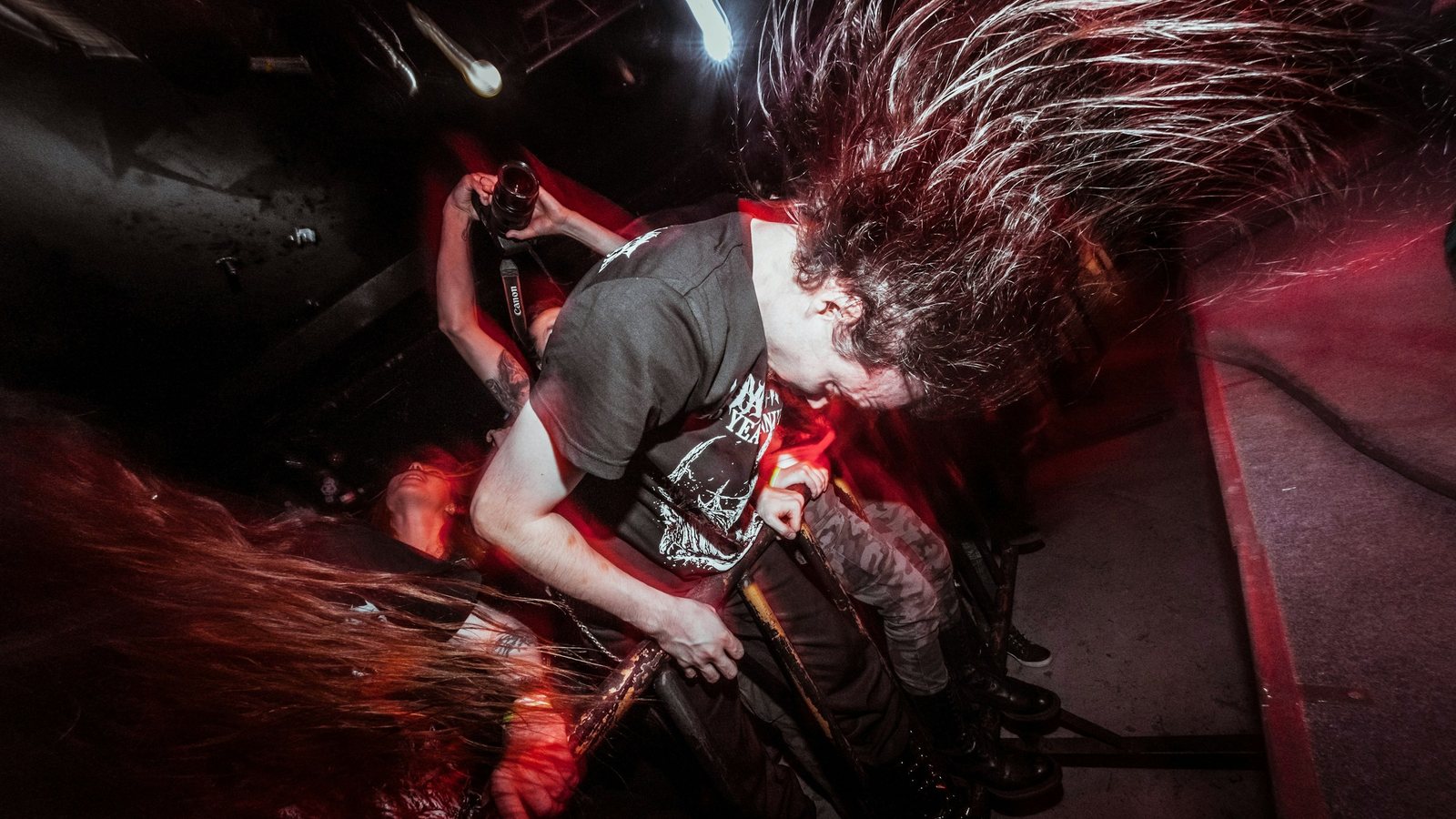 A metal guitarist headbangs on a small club stage, long hair flying in a blur under red light, with another long-haired musician thrashing in the foreground
