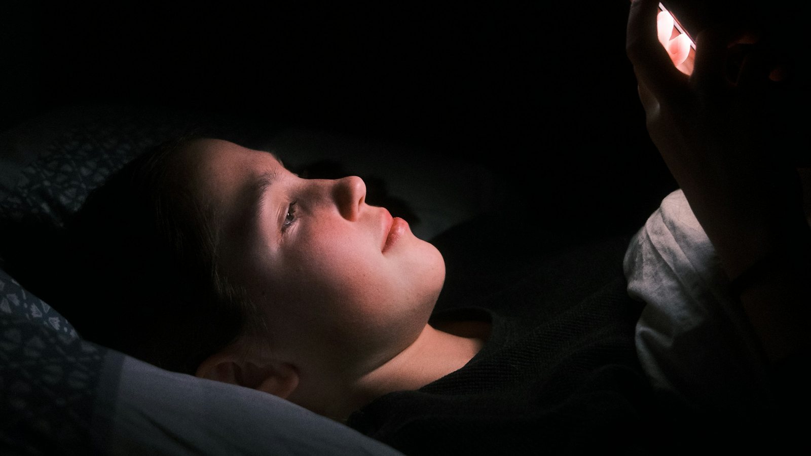 A young woman lies in bed in the dark, her face lit by the glow of the phone she holds above her