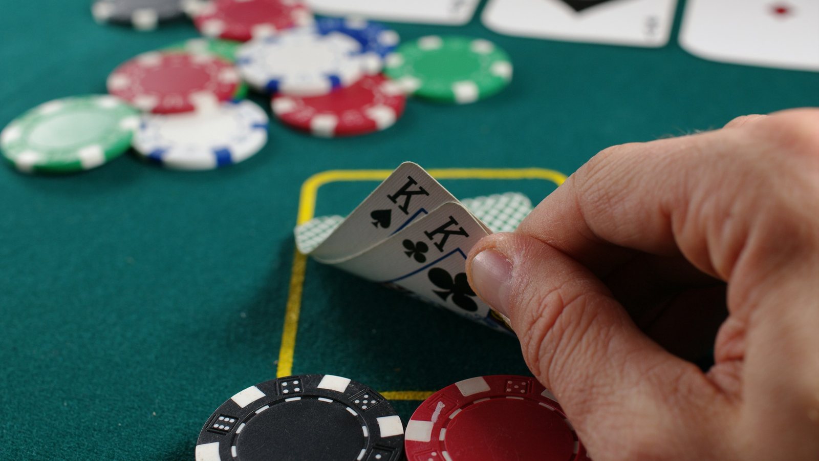 A player lifts the corner of two kings at a green poker table, with stacks of red, black and green chips in front
