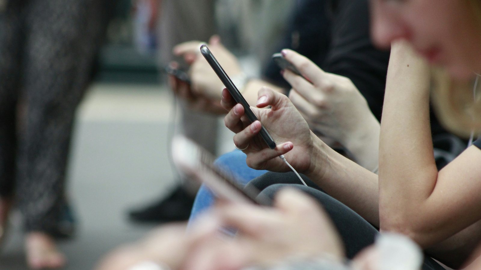 A row of people sitting side by side, each scrolling through a smartphone, with one hand and phone in sharp focus