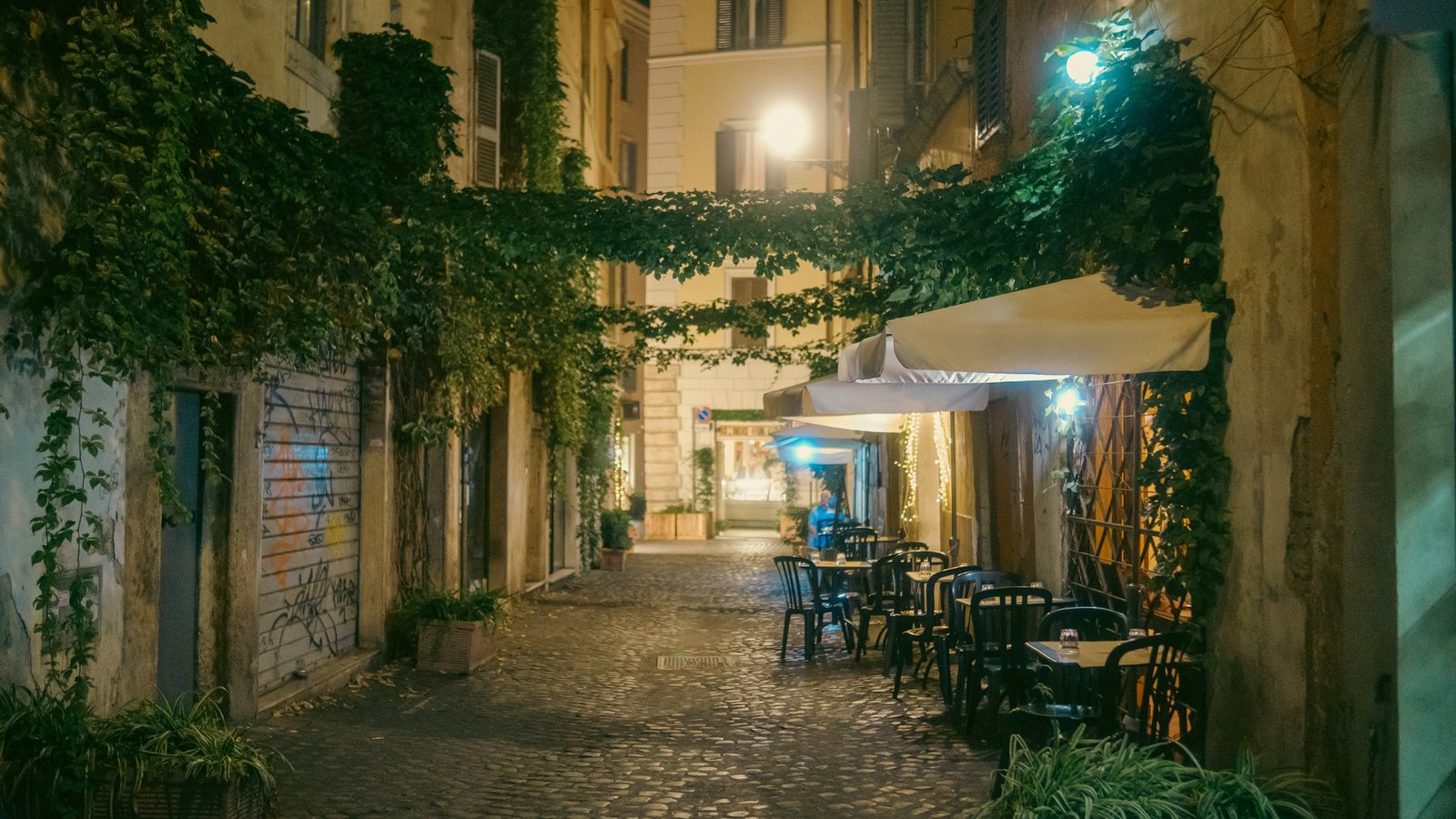 A narrow cobbled street in Rome at night, draped in ivy, with café tables set out under a white awning and warm street lamps