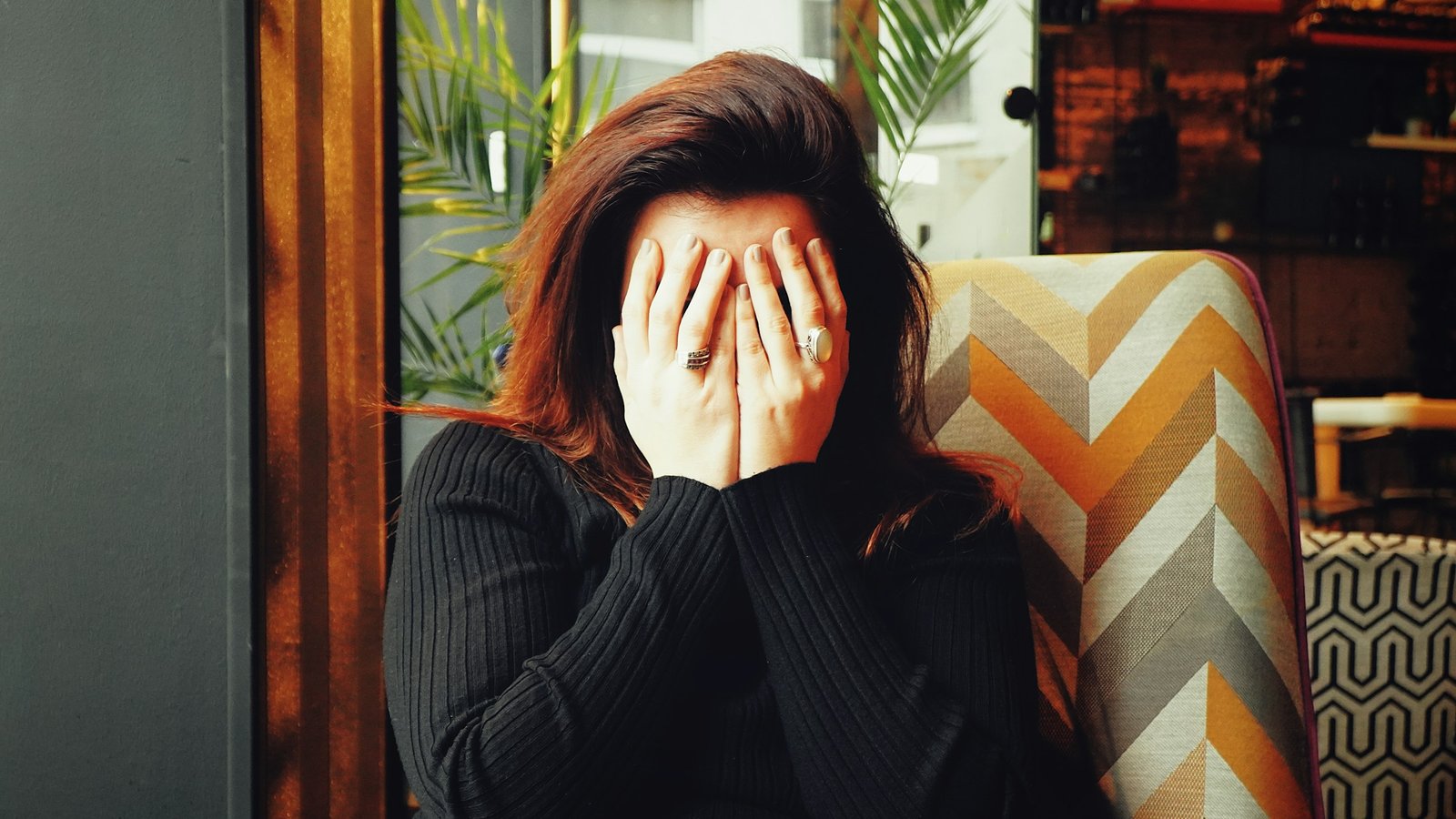 A woman in a black jumper sits in a colourful armchair, hiding her face in both hands