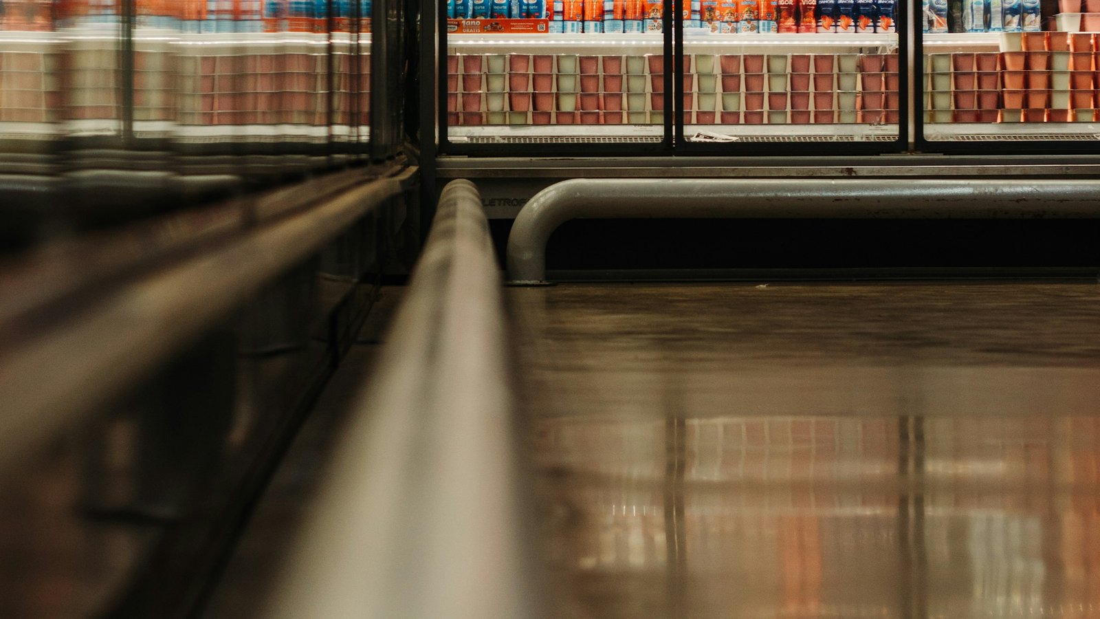 A low view along an empty supermarket aisle at night, chilled-drink fridges glowing behind glass and reflected in the polished floor