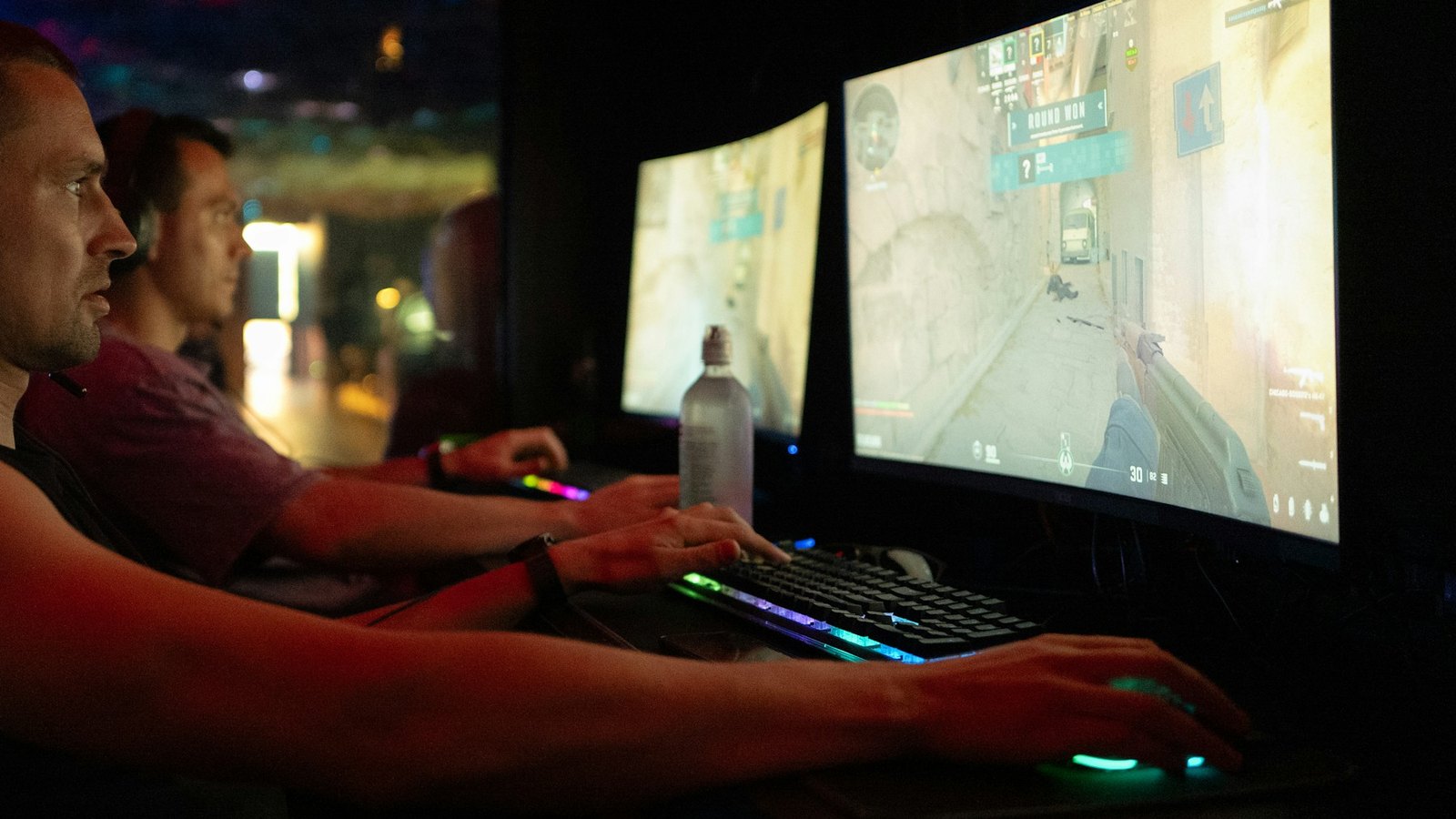 Two players side by side at curved monitors in a dark gaming club, their faces and keyboards lit by the screens