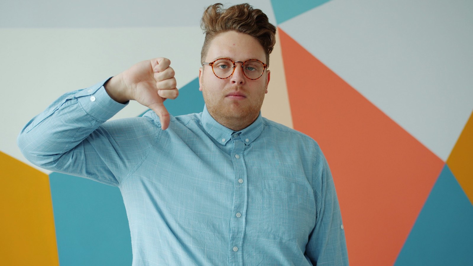 A man in a blue shirt and tortoiseshell glasses holds a thumbs-down gesture towards the camera against a wall of bold colour blocks