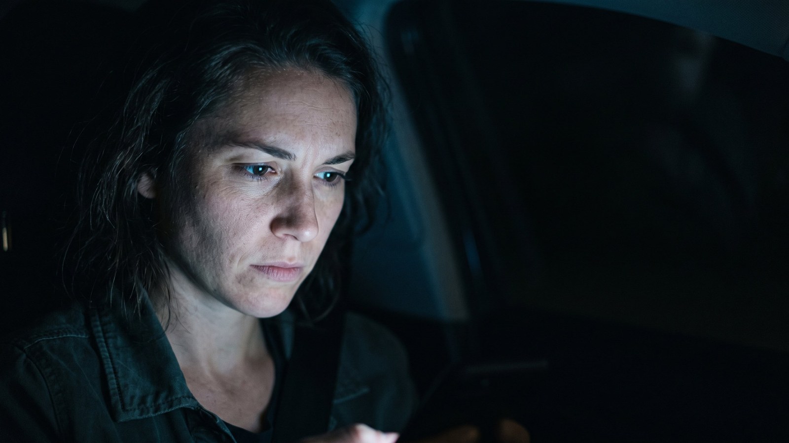 AI-generated image of a woman in a dark room, her face lit by the cold glow of the screen she is studying, wearing a wary, unconvinced expression
