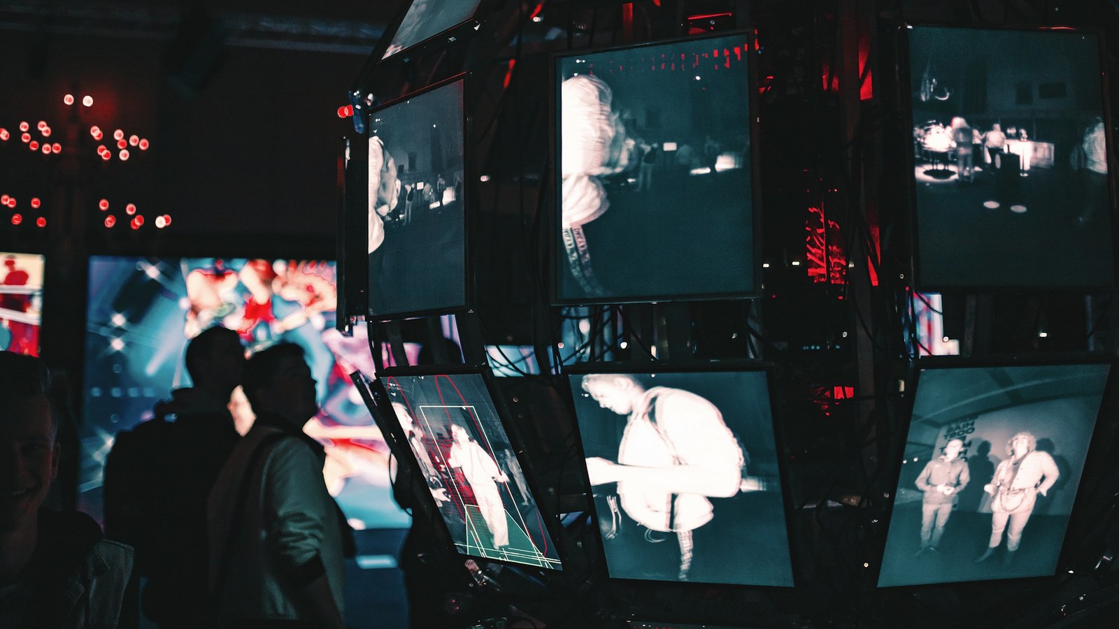 A cluster of screens suspended in a dark hall, each showing a different scene, with people silhouetted watching from below