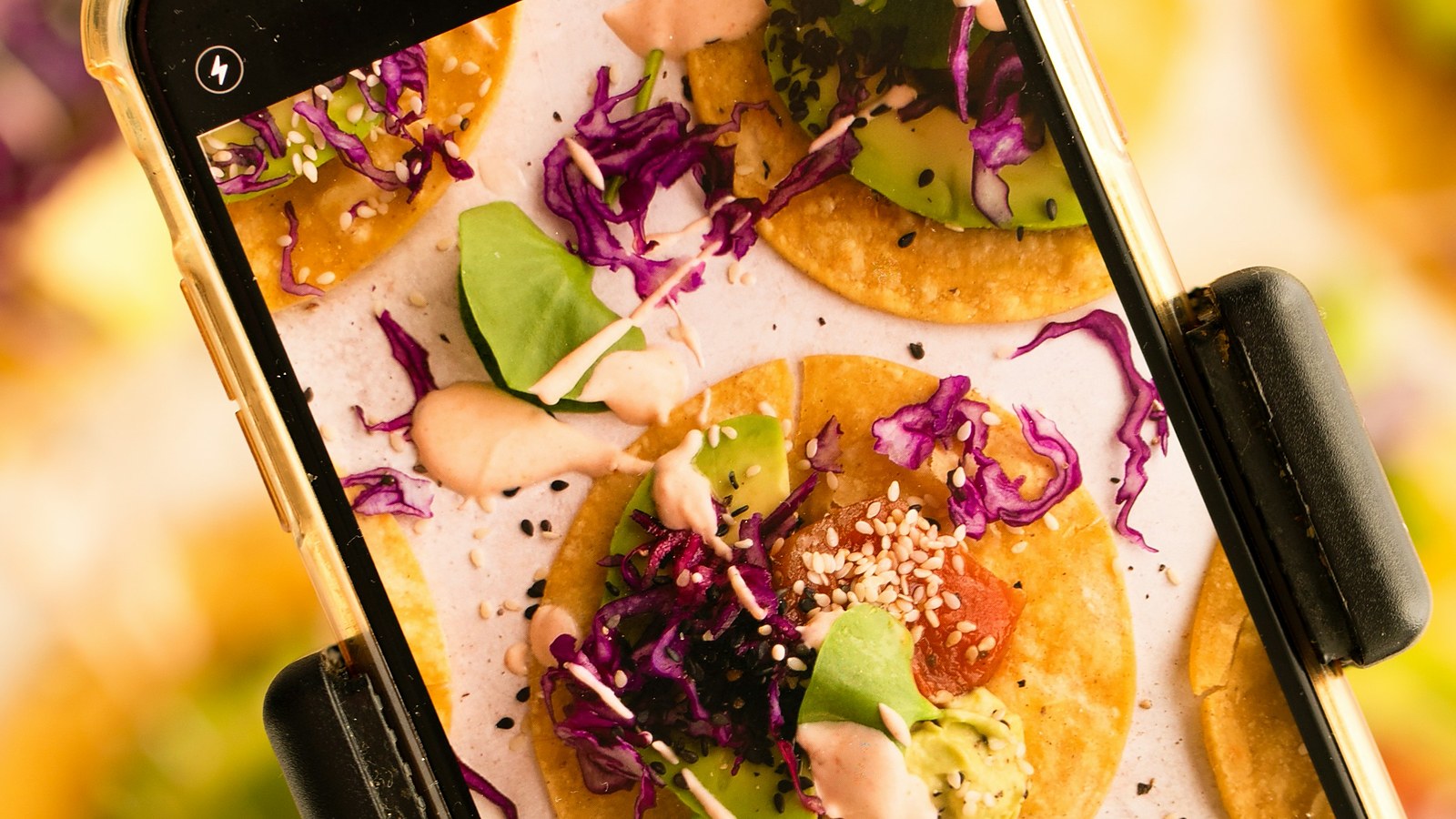 A phone held over a styled plate of tacos, the dish filling the camera screen in sharp colour while the real plate blurs behind it