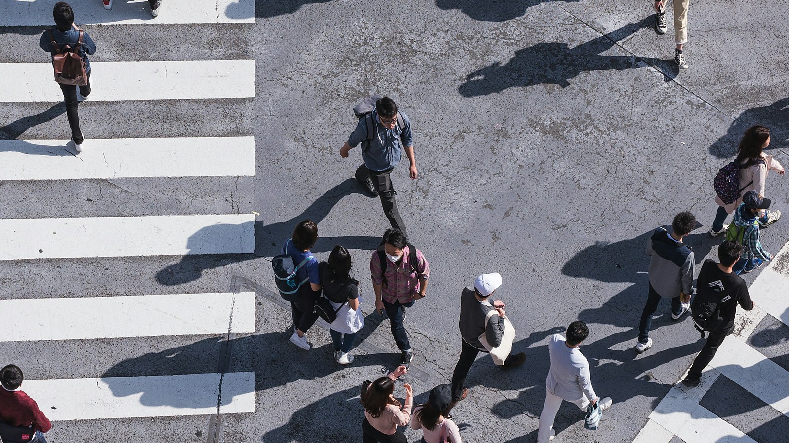 A busy pedestrian crossing photographed from directly above, with dozens of people walking in different directions and long hard shadows across the asphalt