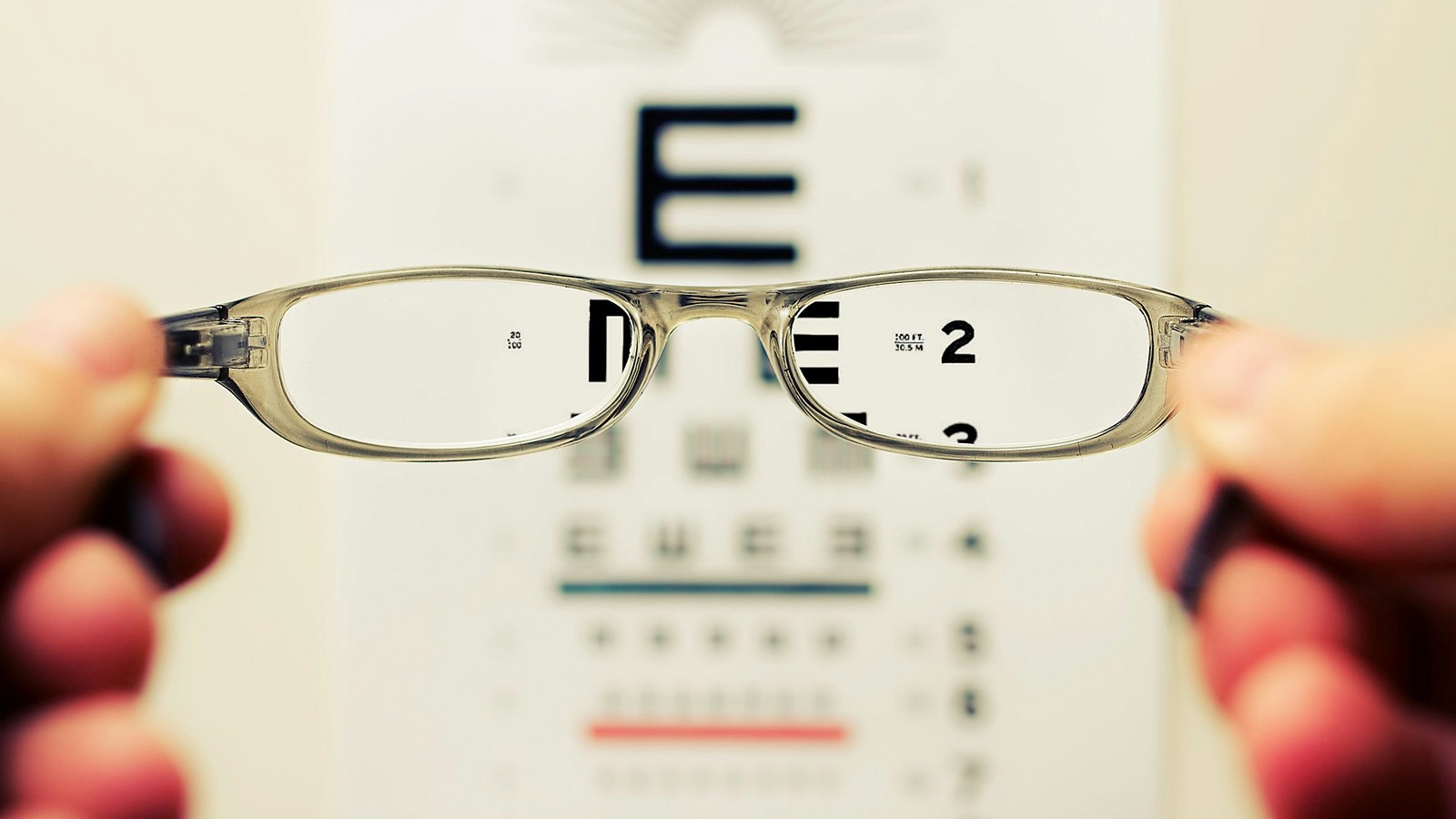 A pair of eyeglasses held up in front of an optician's eye chart, the letters sharp through the lenses and blurred everywhere else
