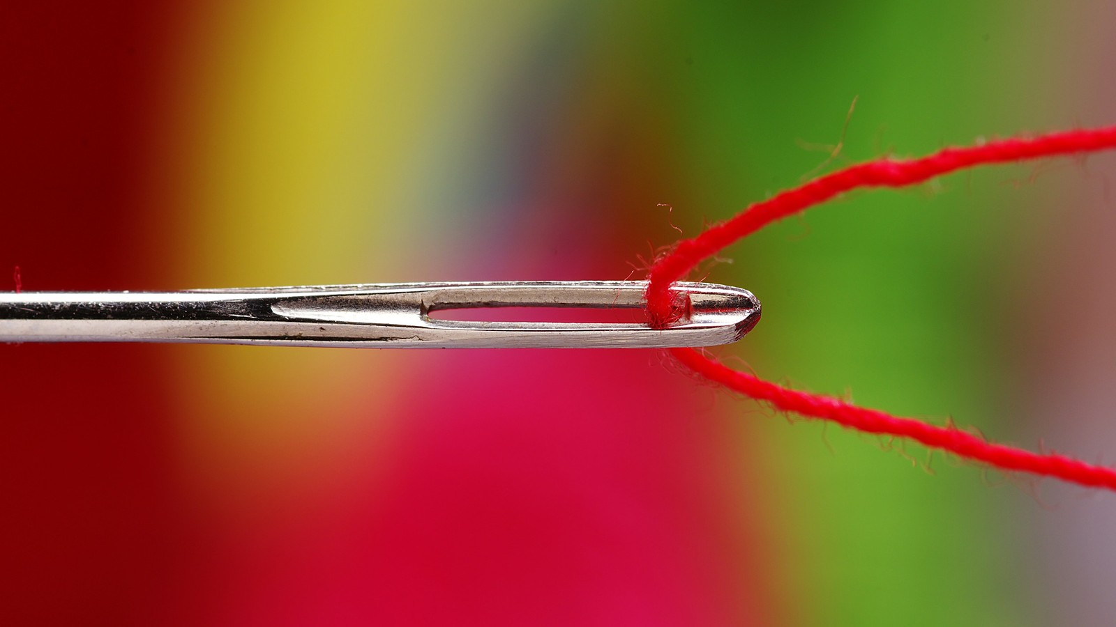 A steel sewing needle in close-up with a bright red thread pulled through its eye, against a blurred background of red, yellow and green
