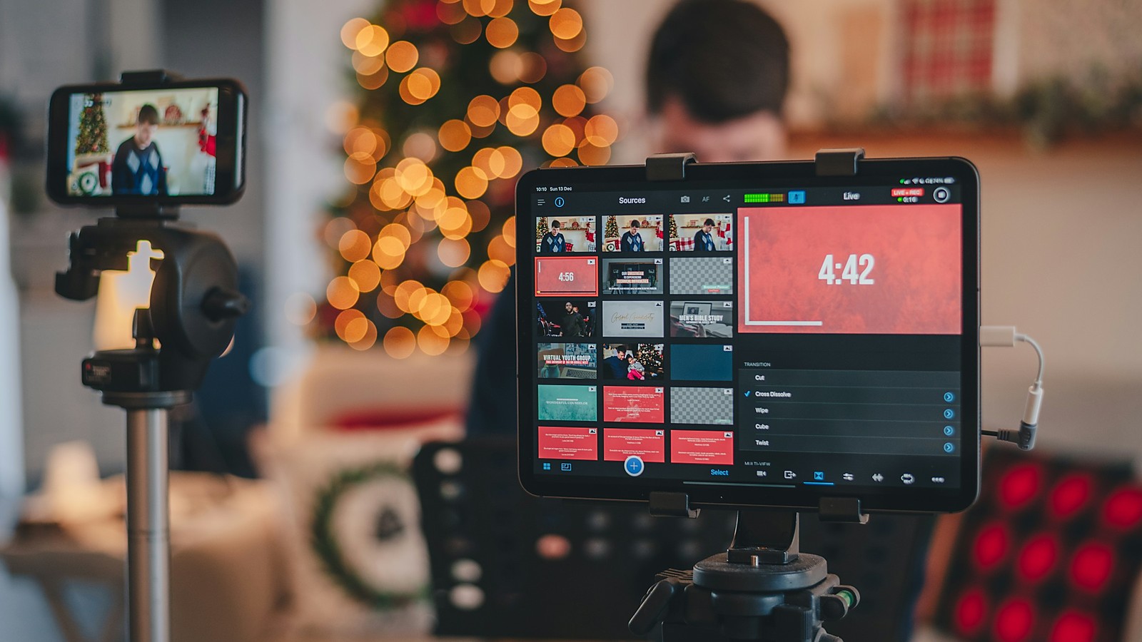 A phone and a tablet on tripods in a living room, the tablet showing a live production dashboard with a timer, a blurred host and warm lights in the background