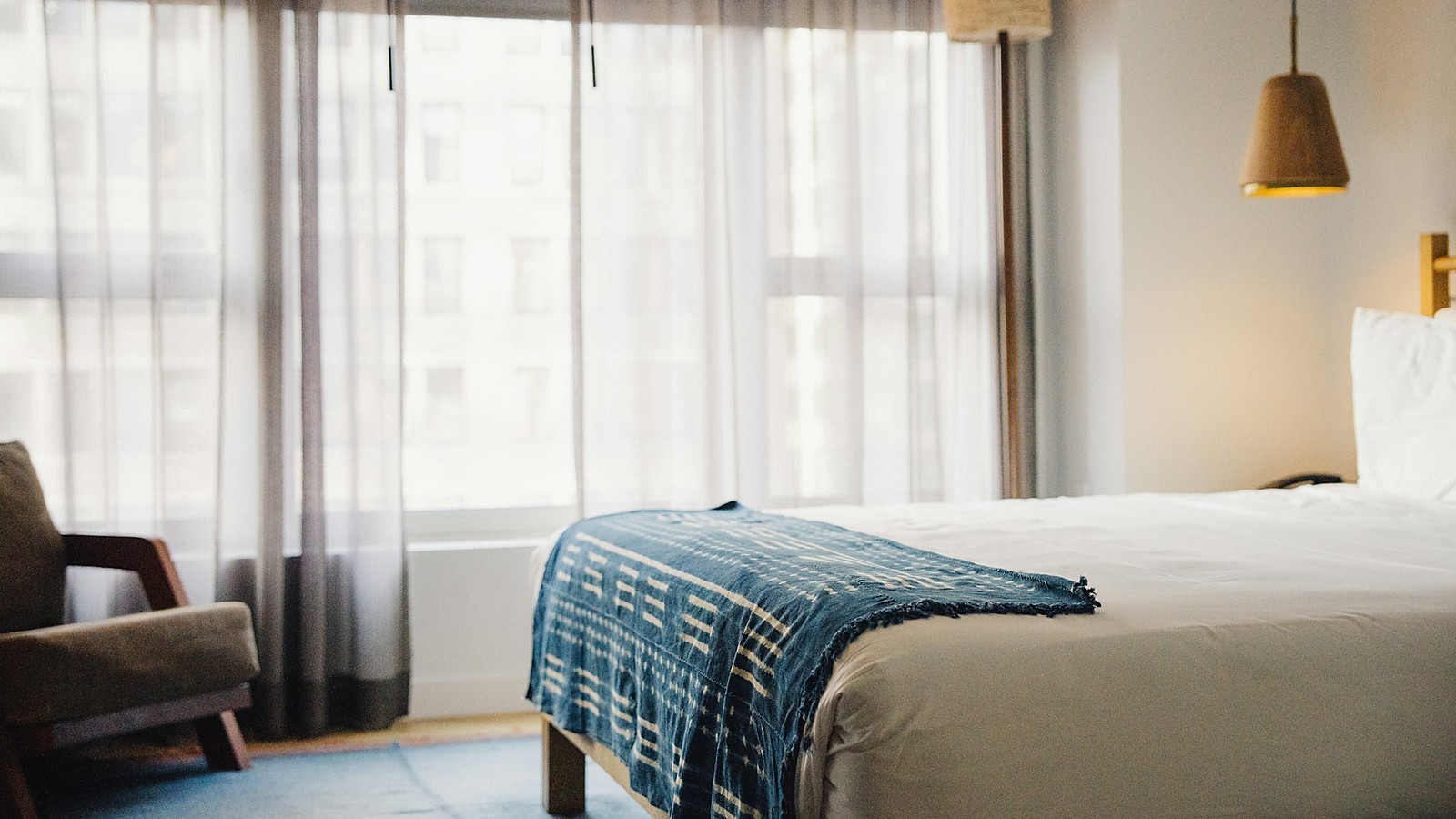 A quiet hotel room with a made bed, a blue throw across the foot, a lamp and an armchair, with soft light coming through sheer curtains