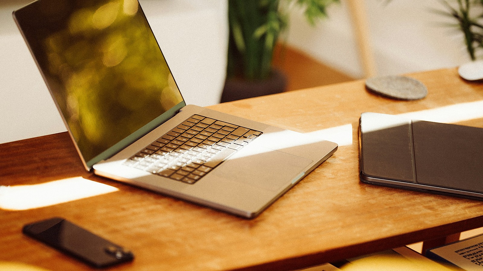 An open laptop on a wooden desk in warm sunlight, with a phone and a closed tablet beside it and a plant behind