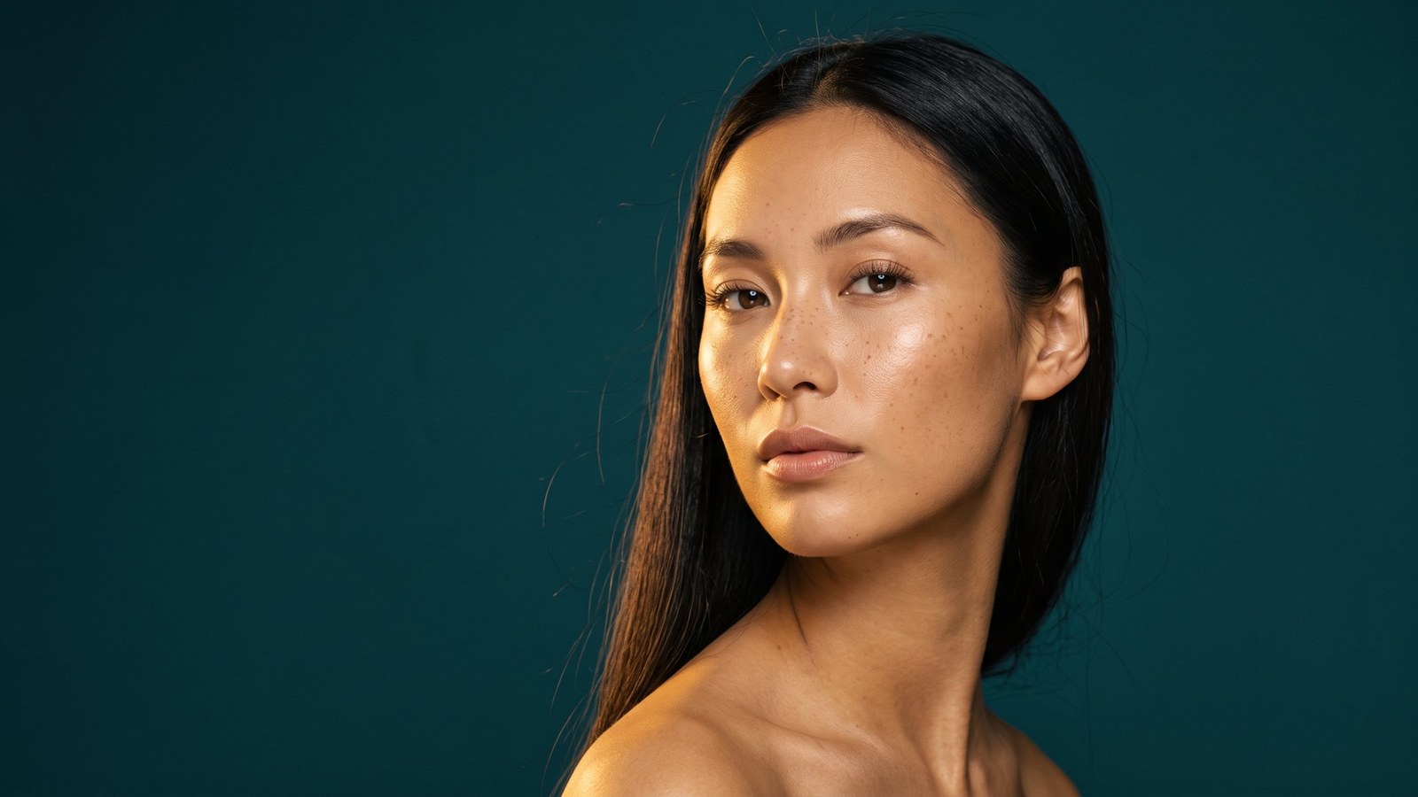 AI-generated beauty portrait of a woman with long dark hair against a deep teal studio background, warm light on her skin, looking straight at the camera