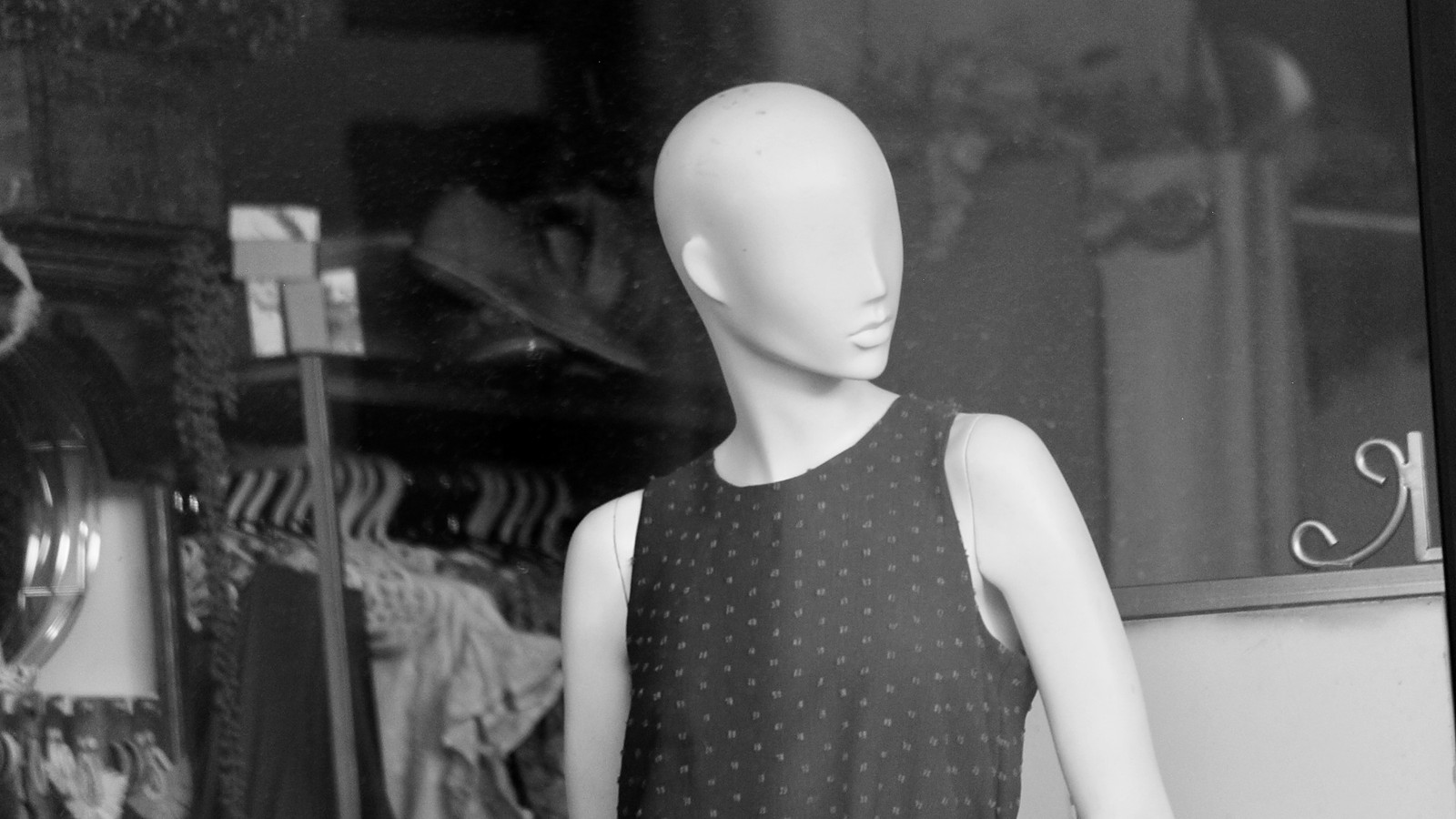 A black and white photo of a shop window mannequin with a smooth, featureless face and a sleeveless patterned top, with clothes on hangers behind