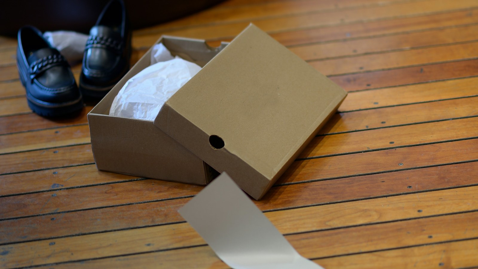 An open cardboard shoe box with tissue paper on a wooden floor, its lid leaning against it, and a pair of black loafers sitting beside it
