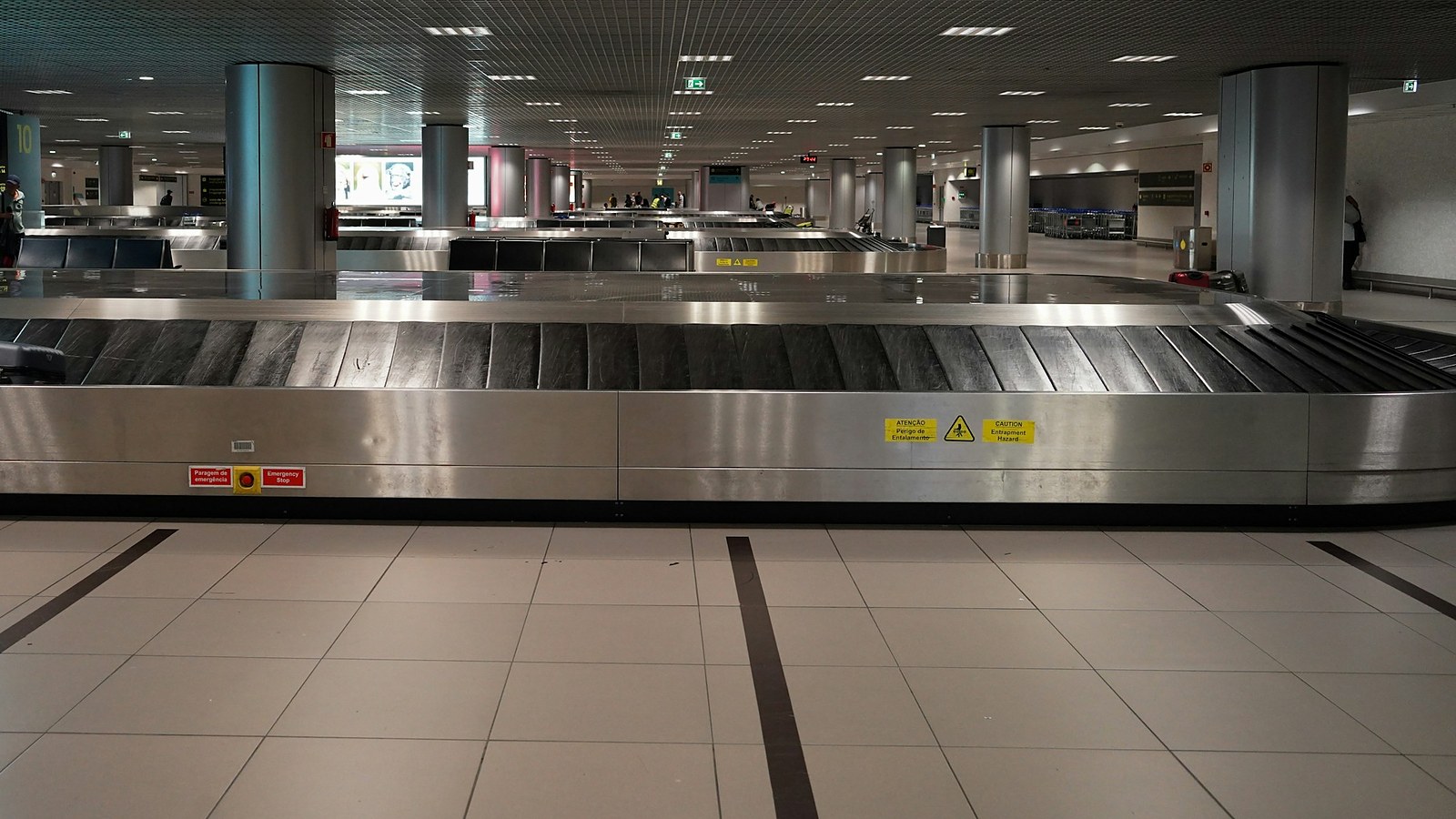 An empty airport baggage carousel with a stainless steel edge and a tiled floor, with more empty carousels stretching into the background