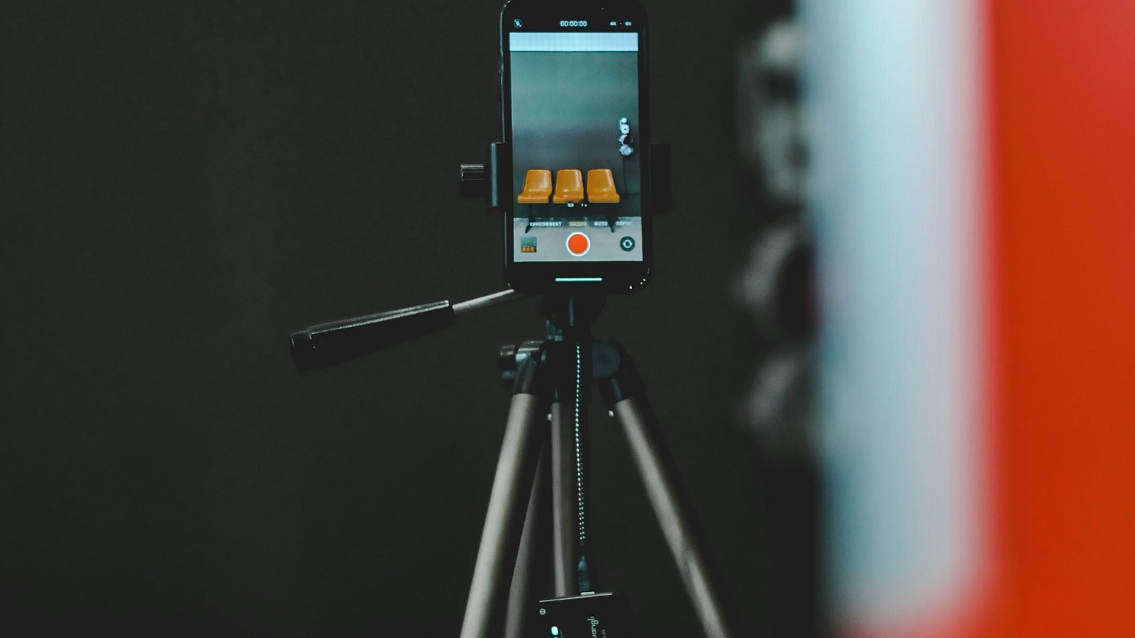 A smartphone on a tripod in a dark studio, its screen showing a camera app ready to record, with a blurred red and white shape in the foreground