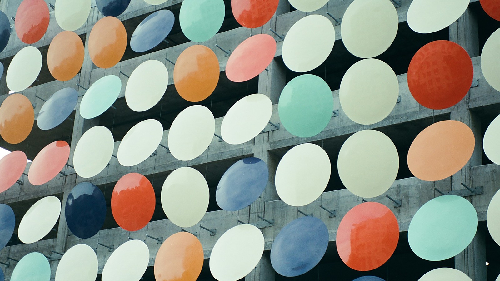 A wall of large oval discs in cream, orange, red, blue and mint hanging in a grid in front of a concrete building